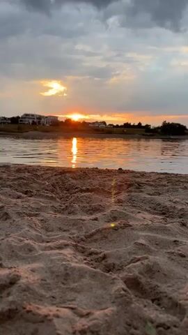 The video shows a close-up view of sand in the foreground, with ripples of water in the middle ground and a sunset in the background. The sky is cloudy, with the sun setting on the horizon, casting an orange glow on the water. There are some buildings visible in the distance. The overall scene is peaceful and serene.