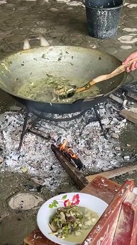 The video shows an overhead shot of a large wok cooking food over an open fire. A wooden spoon is used to stir the food, which appears to be a stew with chunks of fish and green vegetables. Some of the stew is being transferred from the wok to a plate with a floral pattern, which is resting on a red wooden stool. The stool is positioned in front of a wooden chair. The fire is made of logs and ash, and the ground is covered in pebbles and dirt. The lighting suggests it is daytime.