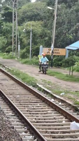 The video shows a railway track in the foreground with a sign in the background that reads "PADHAL" in English and "पांदल" in Hindi. Two people are riding a motorcycle on a dirt path next to the train tracks, approaching the sign. The person driving the motorcycle is wearing an orange shirt and a blue helmet. The person riding behind is wearing a yellow shirt and a white helmet. There are trees and vegetation on either side of the tracks and path. A small covered shelter is visible behind the sign.