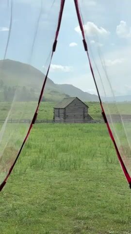 This video shows a view through a tent opening of a wooden structure in a grassy field. The structure appears to be an old log cabin or barn, with a wooden fence in front of it. In the background, there are rolling hills and a blue sky with some clouds. The tent's fabric, a sheer red and white material, is visible in the foreground, framing the scene. The camera is stationary.