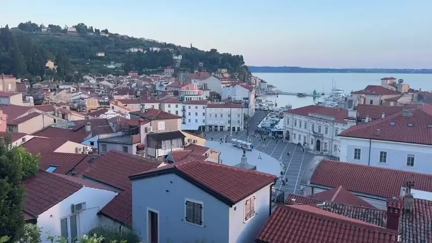 The video shows an aerial view of a European coastal town with red-tiled roofs. The town has a central square with a statue and several people walking around. In the background, there is a harbor with boats and the sea. The sky is a clear blue with some clouds. The architecture suggests an old European town, possibly in the Mediterranean region. The time of day appears to be late afternoon or early evening.