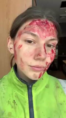 A person is wearing a bright green reflective jacket and has makeup on their face that resembles blood and cuts. The person looks at the camera, then to their left side, and back at the camera. The background shows a door and a kitchen area with white countertops and stainless steel appliances. The lighting is bright and even.