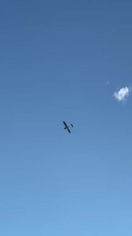 A small airplane with a single propeller is seen flying through a clear blue sky. The airplane is a silhouette against the bright sky, and its wings are spread wide. The video is shot from below, looking up at the airplane. There is a single fluffy cloud visible in the upper right corner of the frame, and another cluster of clouds in the lower left corner. Part of a roof and a satellite dish are visible in the bottom right corner of the frame. The airplane appears to be moving from left to right across the screen.