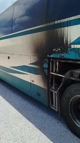 The video shows the exterior of a bus with visible damage from a fire. The bus is parked on an asphalt surface, possibly a roadside. The right side of the bus is prominently displayed, showing sections of the bodywork blackened and scorched. A large tire is visible near the rear of the bus, partially enclosed by a metal grate that appears warped and discolored from heat. The paint on the bus, which is a combination of teal, white, and beige, is peeling and discolored in many areas, with streaks of black soot running down the side. One access panel on the side of the bus is a lighter beige color, but also shows signs of soot and discoloration at its edges. Another section of the bus, to the right of the beige panel, reveals a damaged metal grille with melted or warped horizontal bars, indicating intense heat exposure. The overall impression is of a vehicle that has recently experienced a significant fire, with damage concentrated on its exterior. No people are visible in the video.
