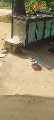The video pans across an outdoor scene. In the foreground, a concrete surface is visible with some debris. A broom is lying on the ground, angled towards the viewer. Moving further back, there are stacks of cardboard boxes, one of which is topped with a clear plastic bag. Adjacent to the boxes is a structure with a metal frame and panels of textured glass, possibly a display case or counter. Inside this structure, a young person is partially visible, looking out. To the right of the structure, on the ground, are various items including what appears to be a tube of sealant, a dark object, and some other unidentifiable items. In the background, there are trees, vegetation, and a dirt path or road. The overall impression is of an outdoor workspace or a makeshift stall.