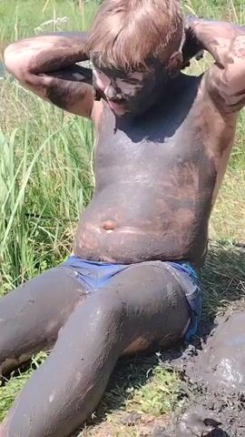 This media file shows a young boy covered in mud, sitting on the ground. He has his hands on the back of his head and appears to be enjoying himself, with a slight smile on his face. The boy is wearing blue swim trunks. The setting appears to be outdoors, with tall grass and reeds in the background, suggesting a natural environment like a riverbank or lake. The lighting indicates it is daytime, likely a sunny day. There is no visible text in the media file. The overall impression is one of playful outdoor activity, possibly swimming or playing in the mud.