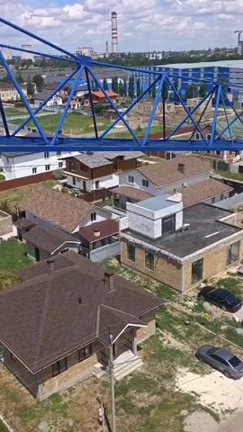 The video shows an aerial view of a construction site. A large blue crane dominates the foreground, its jib extending over a residential area. Below, several houses with orange tiled roofs are visible, some appearing to be under construction. The surrounding landscape is green and hilly, with scattered trees and buildings. The scene suggests ongoing construction activity in a suburban or semi-rural setting. The camera perspective implies it is mounted on a similar structure, possibly another crane or a tall building.