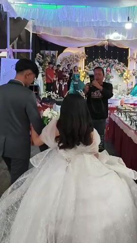 The video shows a wedding reception with a bride in a large white wedding dress and a groom in a dark suit walking towards a decorated stage. Several people are gathered on and around the stage, including a woman in a green dress speaking into a microphone, and other guests. A man in the foreground is holding up a camera or phone, appearing to take a picture or record video. The environment appears to be an indoor venue decorated with white draping, lights, and floral arrangements.