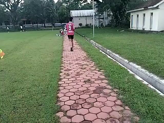 The video captures scenes from an outdoor football (soccer) match, likely in Matesih, Indonesia.

The initial frames show a person wearing a red jersey with the number "9" on the back, running away from the camera on a hexagonal cobblestone path. The path is flanked by green grass fields. In the background, there are trees and several simple buildings with corrugated metal roofs. The sky appears overcast.

The perspective then shifts to show a linesman, dressed in a yellow shirt and black shorts, walking on the grass field while holding a yellow and orange flag. A football goal can be seen further in the background.

The final shot provides a wider view of the playing field, showing multiple individuals engaged in a football game. The field is lush green. In the background, there are various tents and structures, possibly indicating a local event or fair. Distant mountains are visible under a cloudy, late afternoon or early evening sky. The overall setting is an open-air sports ground.
