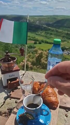A hand stirs a small cup of dark coffee with a spoon. The coffee is in a blue and white patterned cup on a matching saucer. Beside the coffee cup is a clear glass of water, a small antique coffee grinder labeled "Cafe," a small Italian flag on a pole, and a bottle of Doria water. The items are placed on a stone surface. In the background is a lush green valley with rolling hills under a bright blue sky with some clouds. Several coffee beans are scattered on the stone surface. The overall scene appears to be an outdoor setting, possibly a scenic overlook, with an Italian theme suggested by the flag.
