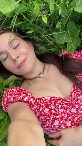 This media file features a young woman lying in a grassy field. She is wearing a red dress with a white floral pattern and a simple necklace with a pendant. Her eyes are closed, and she has a relaxed and peaceful expression on her face. The setting appears to be a natural outdoor environment, possibly a meadow or a field, with lush green grass and other vegetation surrounding her. The lighting suggests it is daytime, with soft natural light. The sky is partially visible in the background, with some clouds, indicating it might be a bright but not intensely sunny day. The presence of strawberry plants with visible strawberries suggests the location might be a strawberry field. The overall mood conveyed is one of serenity and connection with nature. No visible text is present in the media file. The location context is Foteni, Russia.