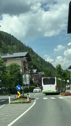 The video begins by showing the rear of a white and green bus driving down a paved road in a town. The bus has a license plate and the text "Salzburg Verkehr" on its back. The road is lined with flower beds and a sidewalk. On the left side of the road, there is a white car driving in the opposite direction, and further down the road, there are more cars. A mountain covered in trees is visible in the background, along with houses and buildings. The sky is blue with fluffy white clouds. The camera appears to be moving, likely from inside another vehicle, as it follows the bus and captures the passing scenery. The video continues to show the bus driving through the town, with other vehicles and pedestrians visible along the roadside. The camera pans slightly to the left, showing more of the street and the surrounding environment. The overall scene depicts a scenic drive through a European town on a sunny day.