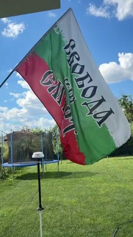 The video shows a Bulgarian flag waving in the wind against a blue sky with scattered clouds. The flag is predominantly white, green, and red, and has black lettering on it. In the background, a green lawn is visible with a trampoline and trees. A small white device on a black pole with wires is in the foreground. The scene appears to be outdoors on a sunny day.
