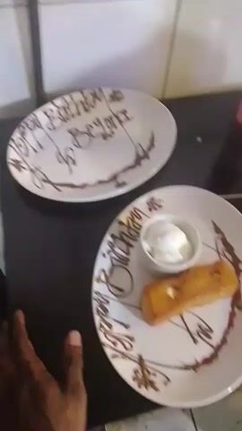 The video shows two white plates on a dark surface. The plate in the foreground contains a rectangular piece of golden-brown pastry, a small white bowl with a white creamy substance, and chocolate drizzle and red sauce decoration. The plate in the background is empty but has a chocolate drizzle decoration and appears to have "Happy Birthday to Beyonce" written on it in chocolate. A hand with dark skin is visible on the left side of the frame, reaching towards the foreground plate. The background is a tiled wall.
