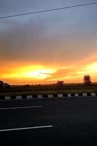 A video captures a dynamic sunset over a highway. The sky is ablaze with vibrant oranges and yellows, gradually transitioning to a dusky blue at the top. Wispy clouds stretch across the horizon, catching the golden light. Below the dramatic sky, a two-lane highway runs horizontally across the frame, its asphalt surface dark and smooth. A white dashed line marks the center of the road, and a white solid line delineates the shoulder. A barrier with alternating black and white stripes separates the highway from the land beyond. Silhouettes of trees and vegetation are visible against the bright sky, hinting at a rural or suburban landscape. Power lines stretch across the upper portion of the frame, adding a man-made element to the natural spectacle. The overall impression is one of a serene and picturesque end to the day, viewed from the perspective of a traveler on the road.