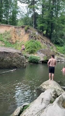 In Irrel, Germany, a group of individuals are enjoying a refreshing dip in a natural body of water. The scene is set amidst a lush, wooded area, with tall trees framing the backdrop and a steep, earthy bank covered in vegetation on one side. Large rocks are scattered around the water's edge, with one prominent boulder serving as a vantage point for a shirtless man standing with his back to the camera, wearing black shorts.

The activity captured is recreational, with one person mid-air, having just jumped or swung into the water from the bank. A splash erupts as they enter the water, creating a dynamic visual. Two other individuals are already in the water, swimming or wading. The water itself appears clear and inviting, reflecting the surrounding greenery. The time of day is difficult to ascertain precisely, but the lighting suggests it could be late morning or afternoon, with the weather appearing to be pleasant enough for outdoor water activities. The overall mood conveyed is one of fun and exhilaration, with the participants embracing the natural environment for leisure. No visible text is present in the media.