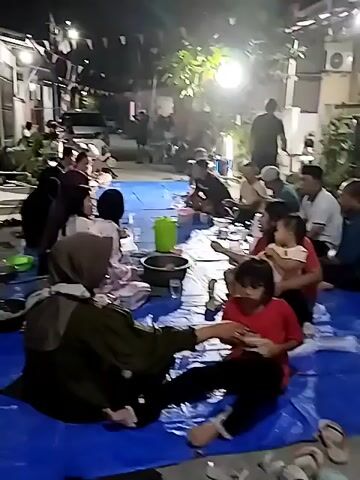 This video shows a nighttime gathering of people, possibly a community event or family meal, taking place outdoors on blue tarps spread across the ground. There are approximately 15-20 individuals visible, including adults and children, seated on the tarps. Food containers and various objects are scattered around them. The environment appears to be a residential area, with buildings and lighting suggesting a street or courtyard. Some individuals are interacting with each other, while others are focused on eating or talking. The lighting is artificial and somewhat dim, creating a warm but low-visibility atmosphere.
