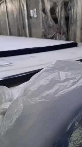 The video shows a sewing machine on a white tabletop. The table is cluttered with tools, including tape, scissors, and a black metal bar. There is a large, flat piece of white fabric on the table, with a dark blue band around its edge. The fabric is also covered in plastic sheeting. A hand can be seen operating the sewing machine, which is cutting the fabric. The background of the video is a cluttered workshop space, covered with plastic sheeting.