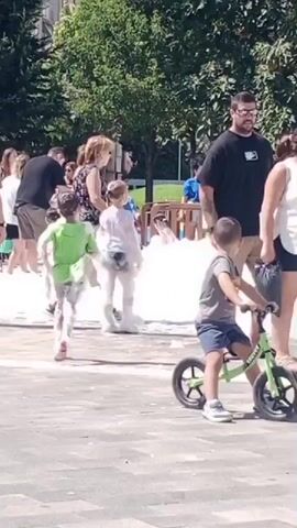 This is a daytime outdoor video capturing a scene with several children and adults. In the foreground, a young boy on a green balance bike is looking towards the left. To the left and slightly behind him, a child in a red swim shirt is facing away from the camera, with soap bubbles on their back and head. Further left, a child in a lime green shirt is running towards the left, also covered in bubbles. In the middle ground, several adults are standing around and observing. One man in a black t-shirt and khaki shorts is looking to his right. A woman in a white tank top and blue shorts is standing next to him. Another woman in a patterned top is talking to a woman in a white t-shirt and jeans. Behind the children and adults, there are trees and some fencing. In the background, more people are visible, and there appears to be a large amount of white foam, possibly from a bubble machine, creating a snowy effect. The ground is paved.