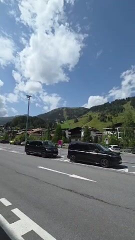 The video shows a paved parking lot with several vehicles, including black minivans and SUVs, parked. The parking lot has white and blue painted lines indicating parking spaces. In the background, there are buildings and trees on a mountainside under a blue sky with white clouds. Several flags are visible, mounted on poles in front of the buildings. A street lamp is also present on the left side of the frame. The perspective is that of someone standing on the pavement, looking towards the parking lot and the mountains.