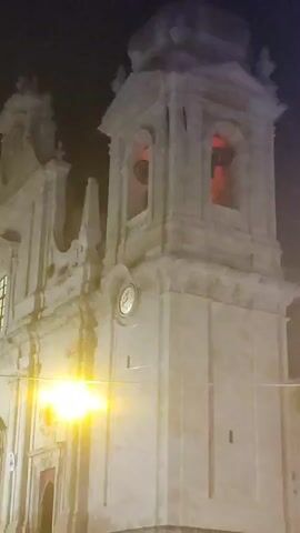 The video shows a nighttime view of a large, ornate church with a prominent bell tower. The church facade is made of light-colored stone and features intricate architectural details, including columns, arches, and statues. The bell tower has openings where bells are visible, illuminated by red light. A clock face is also visible on the tower. Bright yellow lights, possibly streetlights or floodlights, illuminate the front of the church, creating lens flare. Several people are visible on the steps leading up to the church entrance. The sky is dark, indicating it is nighttime.