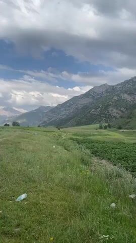 The video shows a panoramic view of a rural landscape with mountains in the background and agricultural fields in the foreground. The sky is overcast with dramatic clouds. The camera pans slowly to the right, revealing more of the expansive scenery. The foreground consists of lush green grass and rows of a cultivated crop, possibly potatoes. In the mid-ground, there are more green fields and scattered trees. The background is dominated by majestic, rugged mountains covered in greenery and rocky outcrops. A dirt path is visible on the right side of the frame. There are no people or animals visible in the video. The overall impression is one of a serene and vast natural environment.