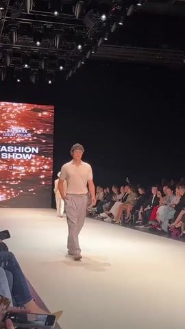 A male model walks down a runway at a fashion show. He is wearing a light-colored short-sleeved polo shirt and light grey pants with a pleated front and a belt. He is also wearing tan sandals. The runway is white and well-lit. To the left of the runway, there is a large screen displaying text that reads "FASHION SHOW". In the background, there is an audience seated on either side of the runway. The lighting is theatrical, with spotlights visible on the ceiling. Another model, wearing a hat and a light-colored outfit, is walking on the runway in the background.