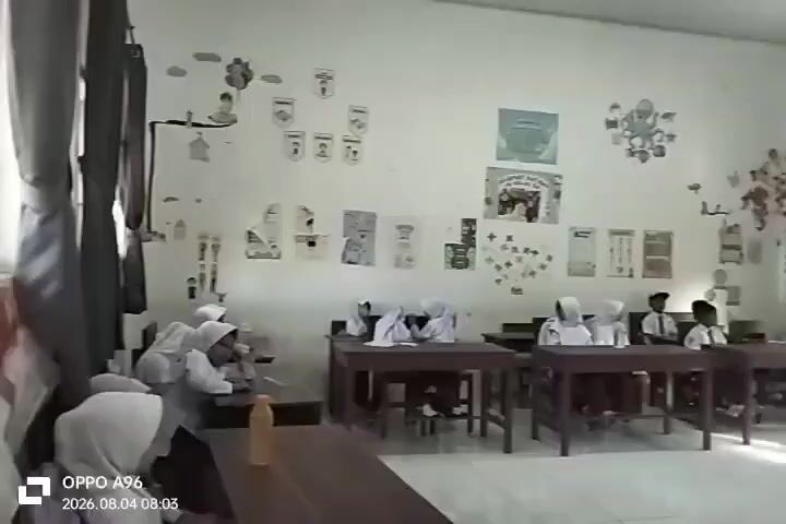 The video depicts a classroom scene with students seated at desks. Several students are wearing white uniforms and headscarves, suggesting they are likely female. The classroom walls are decorated with various posters, drawings, and educational materials, including what appears to be a chart of the alphabet, a poster with cartoon characters, and a drawing of a tree. There are wooden desks and chairs arranged in rows. Sunlight is streaming in through a window, casting shadows on the floor. The overall environment appears to be a typical elementary or middle school classroom. The timestamp at the bottom left corner indicates the video was recorded on August 4, 2026, at 08:03.