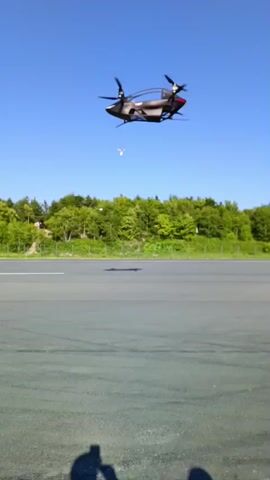 A drone with four propellers hovers in the air against a clear blue sky. It is positioned above a grey asphalt surface, possibly a runway, with a line of green trees in the background. The drone appears to be in flight, with its propellers actively spinning. There is a shadow cast on the ground, suggesting the presence of a person or object.