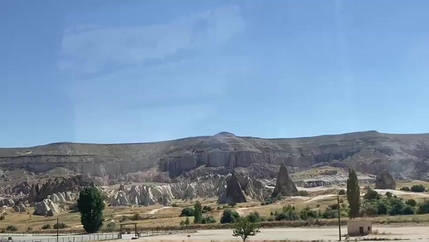 The video shows a wide shot of a desert landscape under a clear blue sky. The terrain is characterized by layered rock formations, some of which are conical in shape. There are sparse patches of dry grass and a few scattered trees. In the foreground, there appears to be a road or pathway with some fencing and what might be picnic tables. The overall impression is of a vast, arid natural environment.