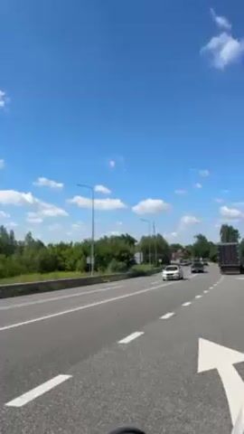 The video shows a road with multiple lanes, with light traffic traveling in the same direction. The sky is blue with scattered white clouds. Initially, several cars are visible, including a white car in the far lane and a dark-colored car further behind. As the video progresses, a white police car with flashing blue and red lights passes by. Following the police car, there is a procession of vehicles including a dark-colored hearse, an olive-green van with a Ukrainian flag on the dashboard, and other cars. A large truck is also visible in the right lane.