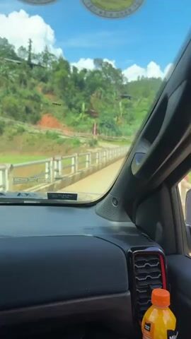 The video shows a car driving over a small concrete bridge. The driver's perspective shows the bridge ahead, framed by the car's dashboard and windshield, the rearview mirror, and part of the driver’s window. On either side of the road, there are green hills and a few buildings. The bridge appears to cross a muddy river. After passing over the bridge, the camera briefly focuses on the river below. The sky is blue, with some scattered white clouds. There is a water bottle on the dashboard.