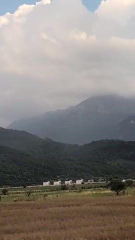 A time-lapse video shows a cloudy sky over a hilly, forested landscape. In the foreground, a field of dry grass transitions to a green field with scattered trees. In the middle ground, a road runs through the fields with a few scattered buildings. In the background, rolling green hills are covered in dense forest. The sky is filled with dramatic, puffy white clouds that change shape and size throughout the short video. The lighting suggests it is either early morning or late afternoon.