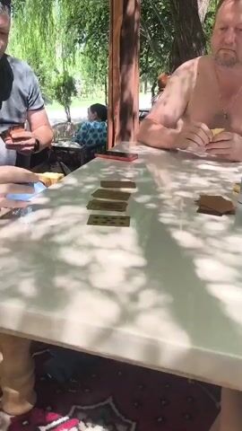 Two men are sitting at a table playing cards, presumably dominoes, outdoors under a shaded structure. The man on the left is wearing a grey t-shirt and is looking at his cards. The man on the right is shirtless and also looking at his cards. There are several dominoes laid out on the table between them, some face up showing dots and others face down. The table is white and the surface is reflective, showing shadows from the overhead trees. There are some containers of spices and a plate with a napkin on the table near the man on the right. In the background, a woman is sitting on a bench and a child is partially visible. The environment appears to be a park or garden setting with lush green trees and foliage.