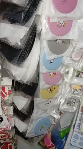 A vertical, close-up, slightly shaky video captures a wall covered in various small, individually packaged items. The items appear to be a mix of personal protective equipment and decorative accessories. On the left side of the frame, there are stacks of black and white face masks, some appearing to be folded and sealed in clear plastic packaging. Interspersed among the masks are small red and white striped stickers.

The right side of the frame displays an array of colorful, circular mirrors or compacts, each individually packaged in clear plastic. These items feature cartoonish designs and come in various pastel colors, including pink, gray, blue, and yellow. Below these, there are rows of stickers in vertical strips, depicting various images such as cartoon characters, food items, clothing, and flowers. Further down, a variety of other small, packaged items are visible, including what appear to be small toys or trinkets in bright, mixed colors. The lighting suggests an indoor environment, possibly a retail display or storage area. No people are visible in the video.