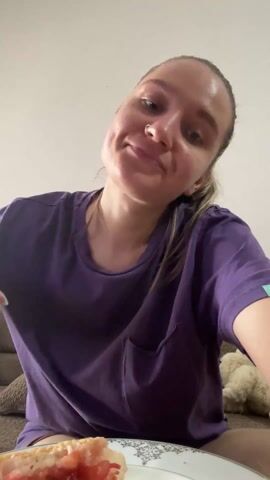 A woman in a purple t-shirt sits on a brown sofa and takes a long drink from a green bottle. A plate with a sandwich sits next to her. The background is a plain, white wall. The woman has blonde hair pulled back in a ponytail. Her left hand is blurred from her movement. The bottle appears to be labeled "SIRMA".