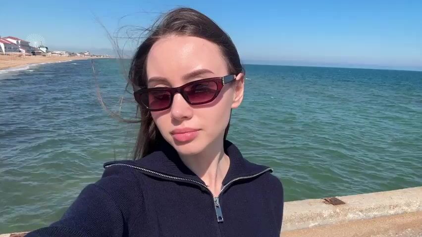 The video shows a woman taking a selfie on a pier. She is wearing sunglasses and a navy blue sweater. The pier is concrete and stretches out into the ocean. The water is turquoise and there are small waves. The sky is blue and clear. In the background, there is a beach with buildings and a Ferris wheel. The wind is blowing and her hair is moving.