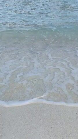 A gentle wave washes onto a sandy beach. The water is clear and shallow, with small whitecaps. The sand is light in color, with visible grains. The scene is calm and peaceful. There are no people or objects visible in the frame. The lighting suggests it is daytime, possibly morning or late afternoon.