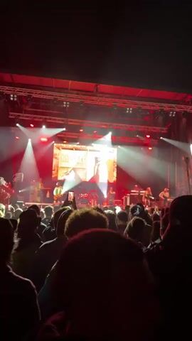 A crowd watches a band called Nine Mile perform on an indoor stage. The lead singer, a woman, is dancing as she sings. Behind the band is a large video screen displaying the band's name and an image of Bob Marley.  The stage is lit with bright white and red lights. Some audience members are recording the performance on their phones.