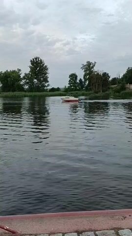A small white and red boat with an outboard motor is seen on a dark, rippling body of water. The boat is positioned in the middle of the frame, heading towards the right. On the left side of the boat, there are tall green reeds and grassy vegetation, with several trees visible behind them. On the right side of the boat, the vegetation continues. In the background, the sky is overcast with gray clouds. The foreground shows the edge of a dock or walkway with pink and white cobblestones and a red railing. The water is reflecting the trees and the sky.