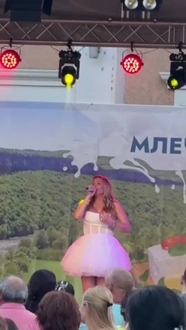 A woman with blonde hair is singing into a microphone on a stage. She is wearing a white top and a pink tutu skirt. There are lights above her, some red and some blue. Behind her is a large banner with a picture of trees and the word "МЛЕЧ" visible. There are people in the audience in front of the stage, some of whom are looking at the singer.