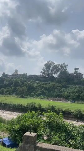 A person is filming from a moving vehicle. The camera pans to the left showing a lush green hillside dotted with trees and some buildings. Below the hill is a large, green field, possibly a sports ground. A dry riverbed with rocks is visible in the foreground. The sky is cloudy with patches of blue.