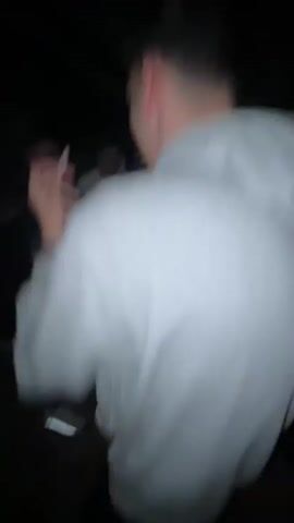 The video appears to be taken inside a room at night, possibly at a party or gathering. Initially, the view is focused on the back of a person wearing a white hoodie. The camera then moves to show a group of people around a table with various items on it. The perspective shifts again to focus on a person lounging in a chair, holding up a phone to take a photo or video. The scene is dimly lit and the details are somewhat blurry due to the low light conditions.
