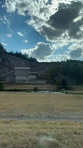 The video shows a view from a moving vehicle, looking out a side window. The scene is a rural landscape with a large industrial building, possibly a quarry or processing plant, situated at the base of a rocky, tree-covered mountain. In the foreground, there is a dry, grassy field. A river or stream is visible between the field and the industrial building. The sky is blue with scattered white clouds. A side mirror of the vehicle is visible in the lower portion of the frame. The video appears to be taken during daylight hours.