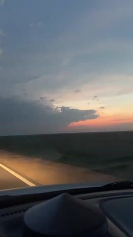 This media file shows a view from inside a vehicle driving on a highway at dusk. The setting is a flat, open landscape with fields on either side of the road. In the distance, several wind turbines are visible, suggesting a rural or open area. The sky is filled with dramatic clouds, with a sliver of orange and pink sunset peeking through the darker grey clouds on the horizon.

The activity is driving on a highway. The headlights of the vehicle are on, illuminating the road ahead, and the road has a bright yellow line marking the lane. Other vehicles are visible on the highway, with their headlights on, indicating low visibility due to the time of day. The overall mood is peaceful and slightly melancholic, evoked by the dimming light and the vast, open landscape. The time of day is clearly dusk, transitioning into night. There is no visible text in the media file.