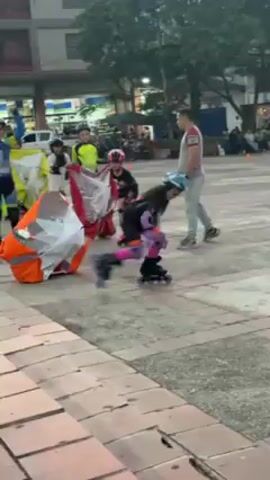 The video captures a person on roller skates, being pulled by a large orange windsock. The skater, wearing a helmet, knee pads, and purple gloves, navigates around a course marked by green and yellow cones on a tiled plaza. The orange windsock inflates and deflates as it is pulled, creating drag and resistance for the skater. In the background, there are several people seated on benches watching the activity. Additional individuals are visible in the distance, along with trees and buildings under a bright sky, indicating an outdoor setting. The camera is at ground level and follows the skater's progress throughout the course.