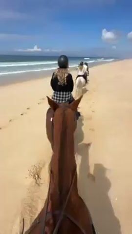 The video shows a group of people horseback riding on a sandy beach. The sky is partly cloudy and the ocean is visible with small waves. The person recording the video is on a brown horse and turns the camera around to show their face, smiling. The video then pans back to the view of the beach.