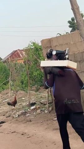 A person walks from left to right in a side profile, carrying a sewing machine on their shoulders. The sewing machine is black and white and sits on a light-colored base. The person is wearing a dark blue shirt with some writing on the back and black pants. They have yellow sandals on their feet. The environment is a dirt path with some vegetation and buildings in the background. A rooster is visible in the left background. The sky is overcast.
