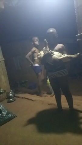 The video shows four young boys in a dimly lit room with a dirt floor. One boy holds a white rooster, another stands beside him, and two other boys are standing nearby. There is a bag labeled "Sleeping" and some metal bowls in the lower left corner. A blue plastic chair is visible in the background. The scene appears to be indoors, possibly in a home or shelter. The light source is coming from above, creating strong shadows.