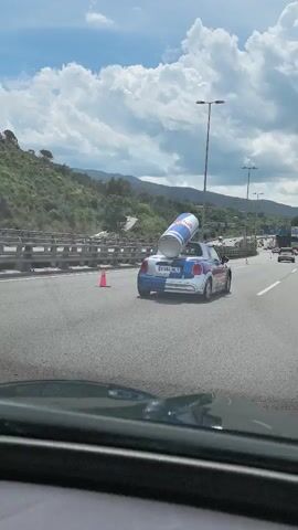 The video shows a vehicle driving on a multi-lane highway during the daytime. The sky is partly cloudy. The vehicle in the foreground is following a blue and white Mini Cooper with a large Red Bull can mounted on its rear. The Mini Cooper is traveling in the lane to the right of the vehicle recording the video. Other vehicles are visible in the distance. The side of a hill is visible to the left of the highway. Orange traffic cones are also visible along the side of the highway.